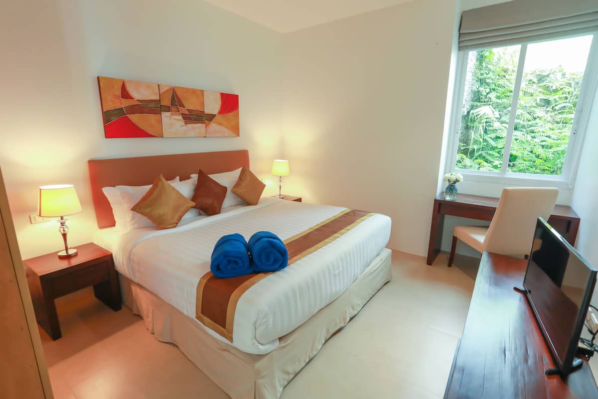 A hotel room with a large bed, two bedside lamps, a desk with a chair, and a window with a view of greenery.