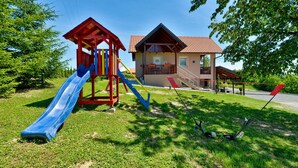 Ein Spielplatz mit Rutsche und Klettergerüst, ein Haus mit rotem Dach und ein klarer, blauer Himmel.