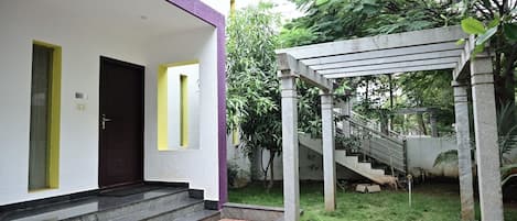 Un edificio con fachada morada y blanca, una escalera que lleva a un espacio cubierto y un pequeño jardín con césped y árboles.