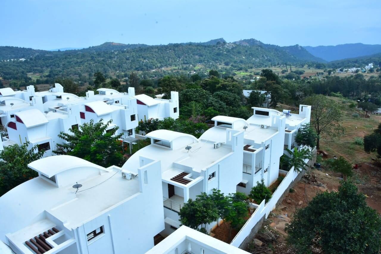 A residential area with white buildings, some with flat roofs and others with sloped roofs, surrounded by greenery and hills in the background.