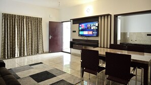 A modern living room with a flat-screen TV, a dining area with a table and chairs, a ceiling fan, and a large window with curtains. - Ags Holiday Resorts (Tirupattur)