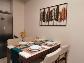 Uma área de refeições com uma mesa de madeira posta para quatro pessoas, uma peça decorativa acima e um frigorífico à esquerda.