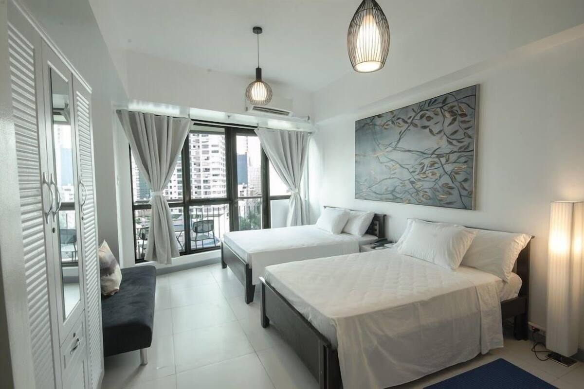 A modern hotel room with two beds, a large window with curtains, a ceiling fan, and a wall-mounted artwork.