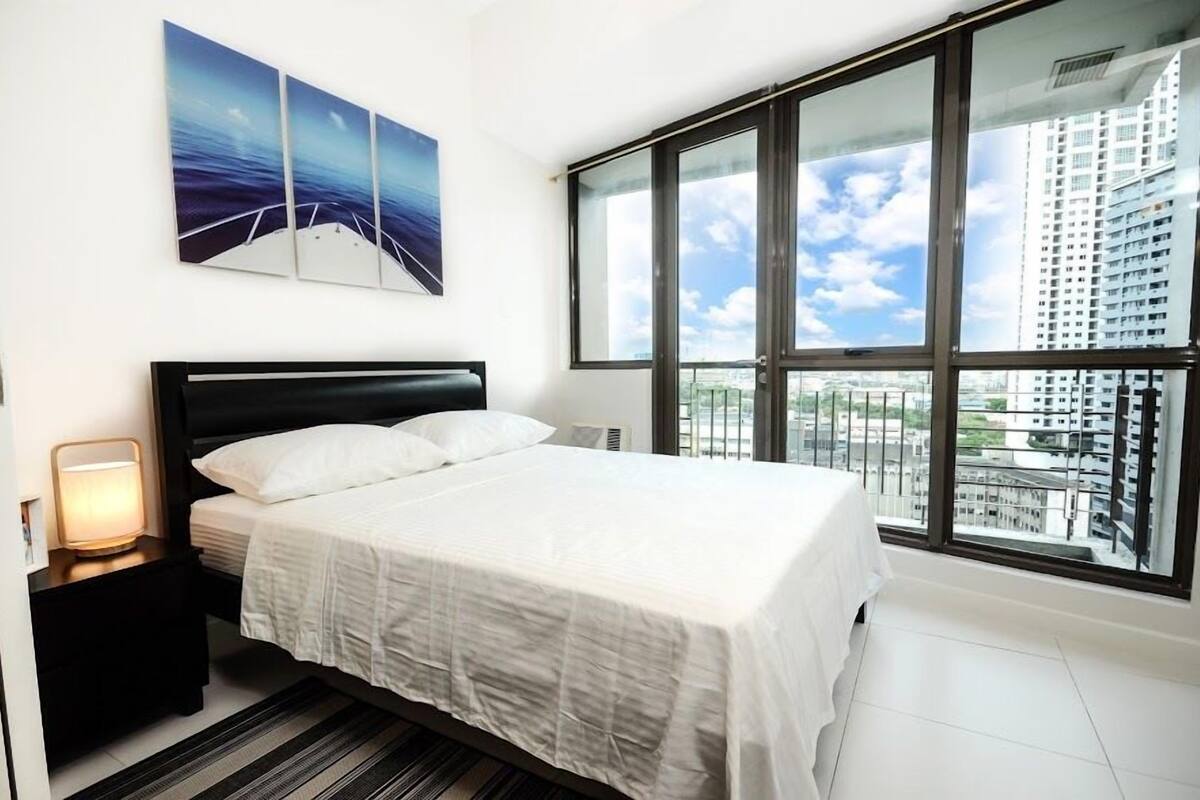 A modern hotel room with a large bed, a nightstand with a lamp, a view of the city through large windows, and a painting on the wall.