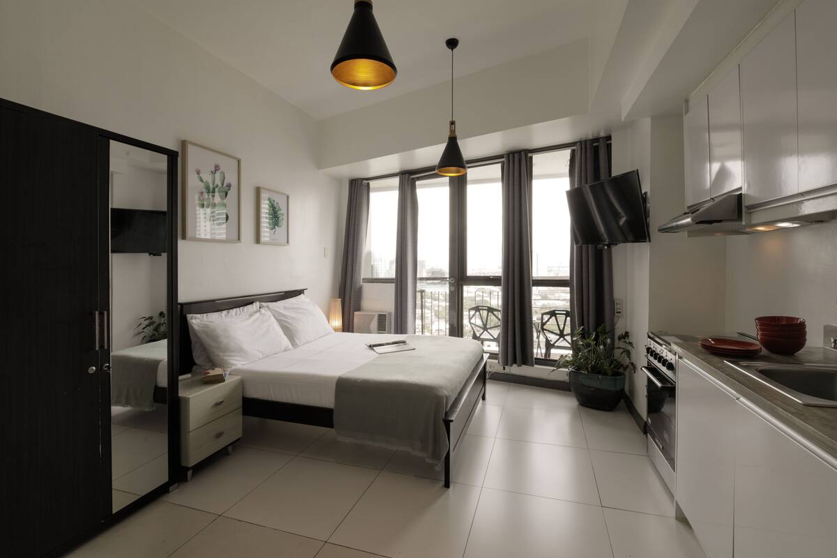 A modern bedroom with a bed, bedside tables, a nightstand, a dresser, a TV, and a large window with curtains.
