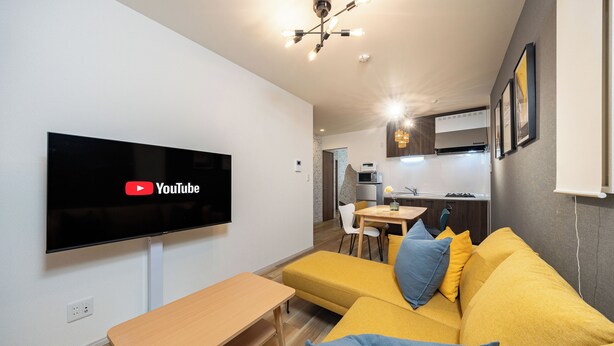 A modern living room with a flat-screen TV, a yellow sofa, a wooden table, and a kitchen in the background.