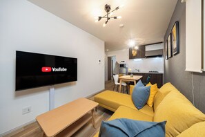 A modern living room with a flat-screen TV, a yellow sofa, a wooden table, and a kitchen in the background. - Waraku (Osaka)