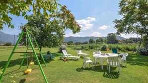 A grassy outdoor area with a swing set, white chairs, and a table with cups, surrounded by trees and a view of hills.