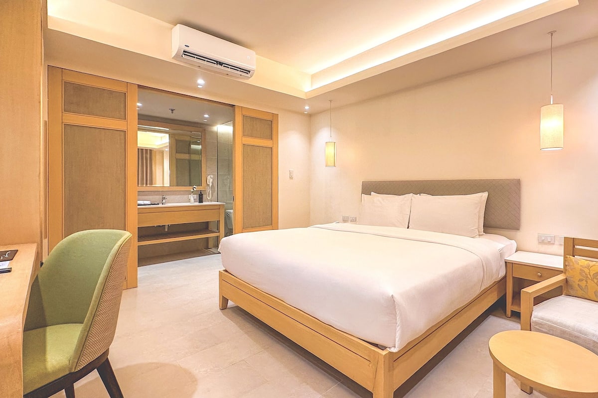 Habitación de hotel con cama, escritorio, silla, mesita de noche, espejo y aire acondicionado.