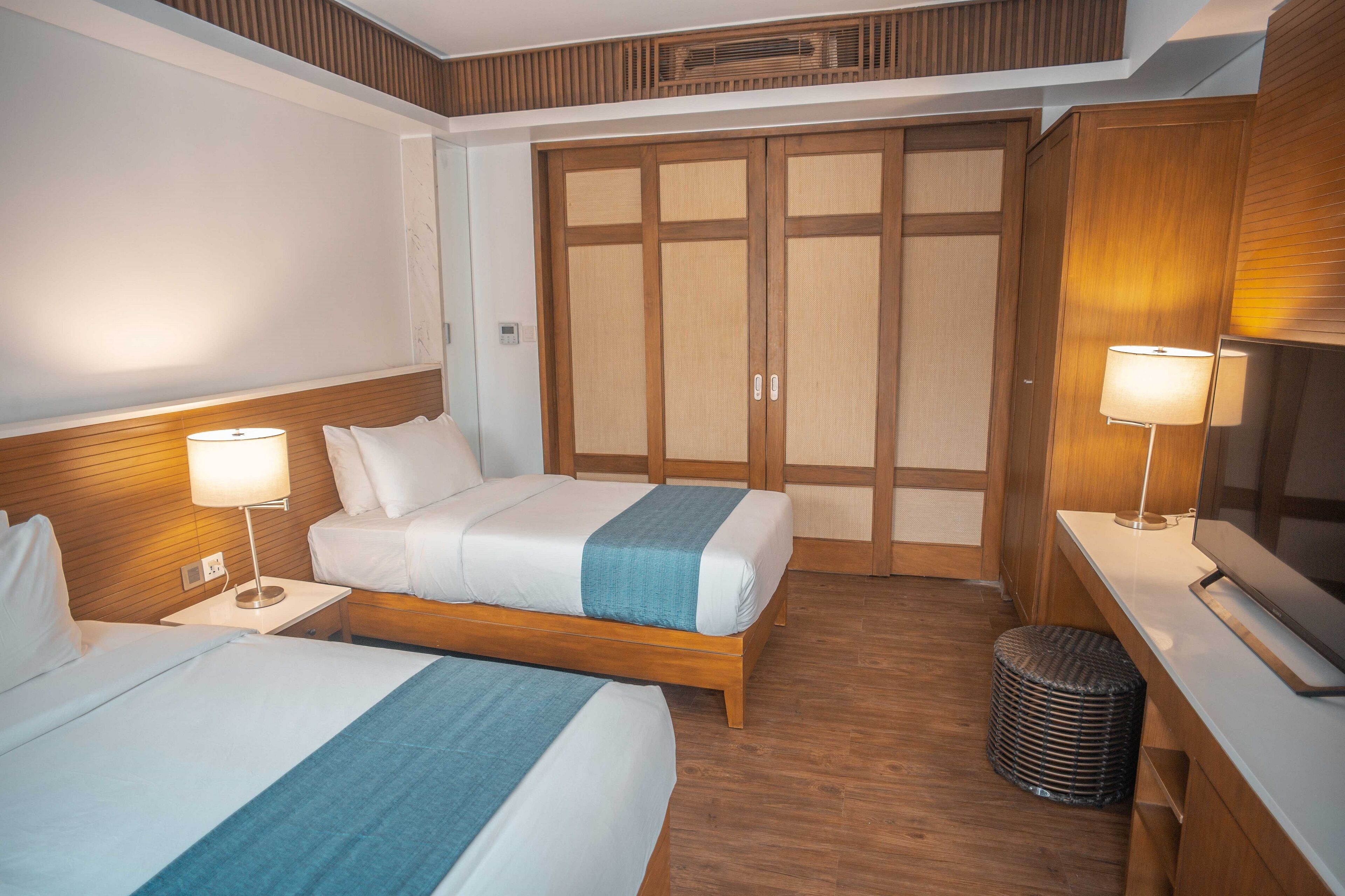 Ein Hotelzimmer mit zwei Betten, Holz-Kopfteilen, einem großen Kleiderschrank, einer kleinen Mülltonne und einem Flachbildfernseher.