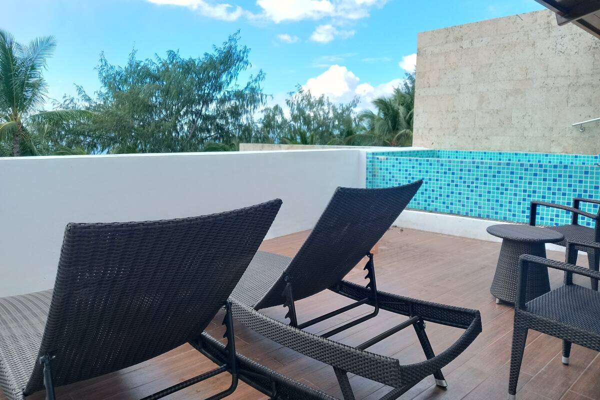 Un patio con sillas de mimbre y una pequeña mesa, con vista a una piscina con baldosas azules.