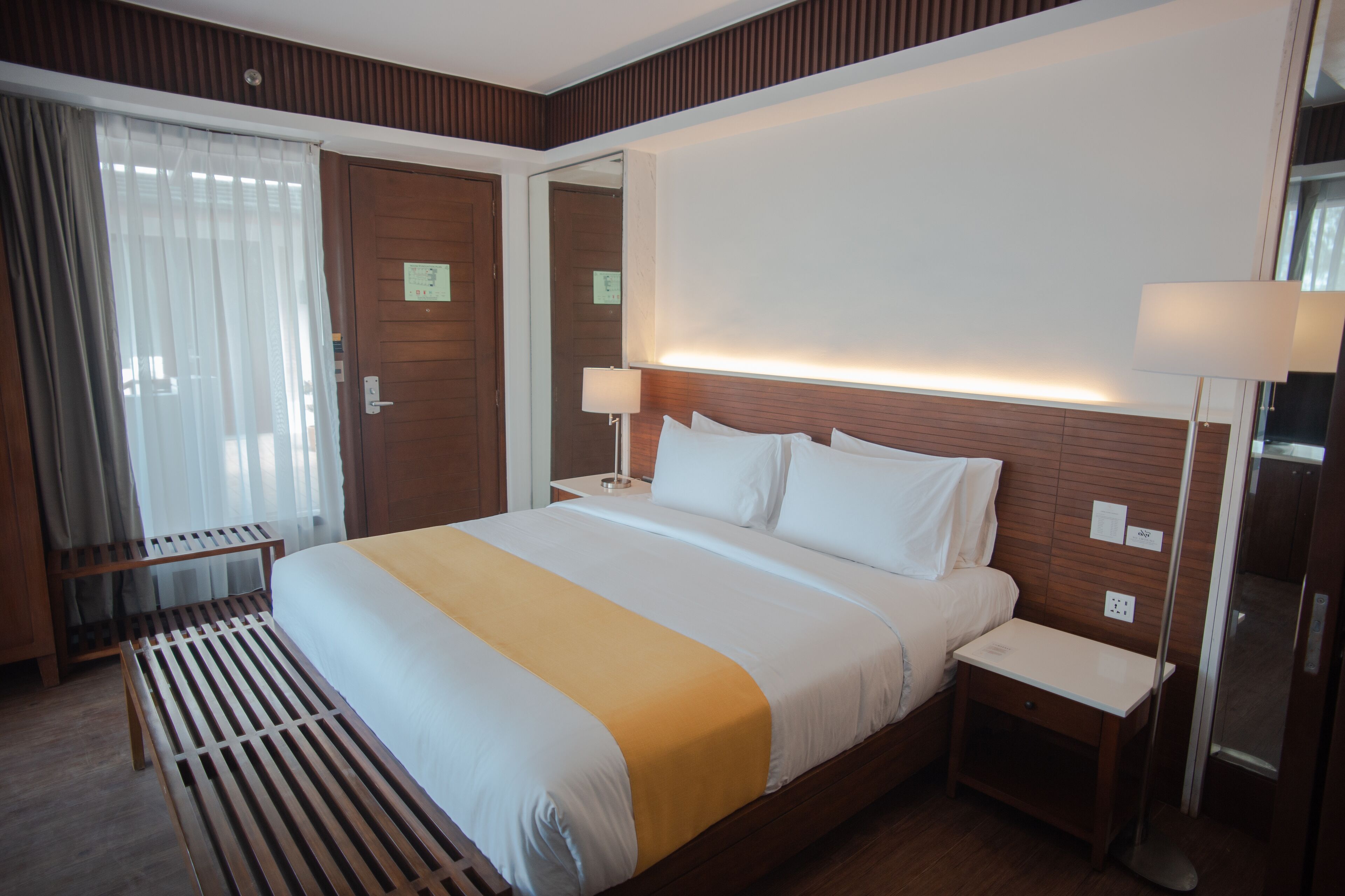 A hotel room with a large bed, wooden headboard, bedside tables, a bench, and a mirror.