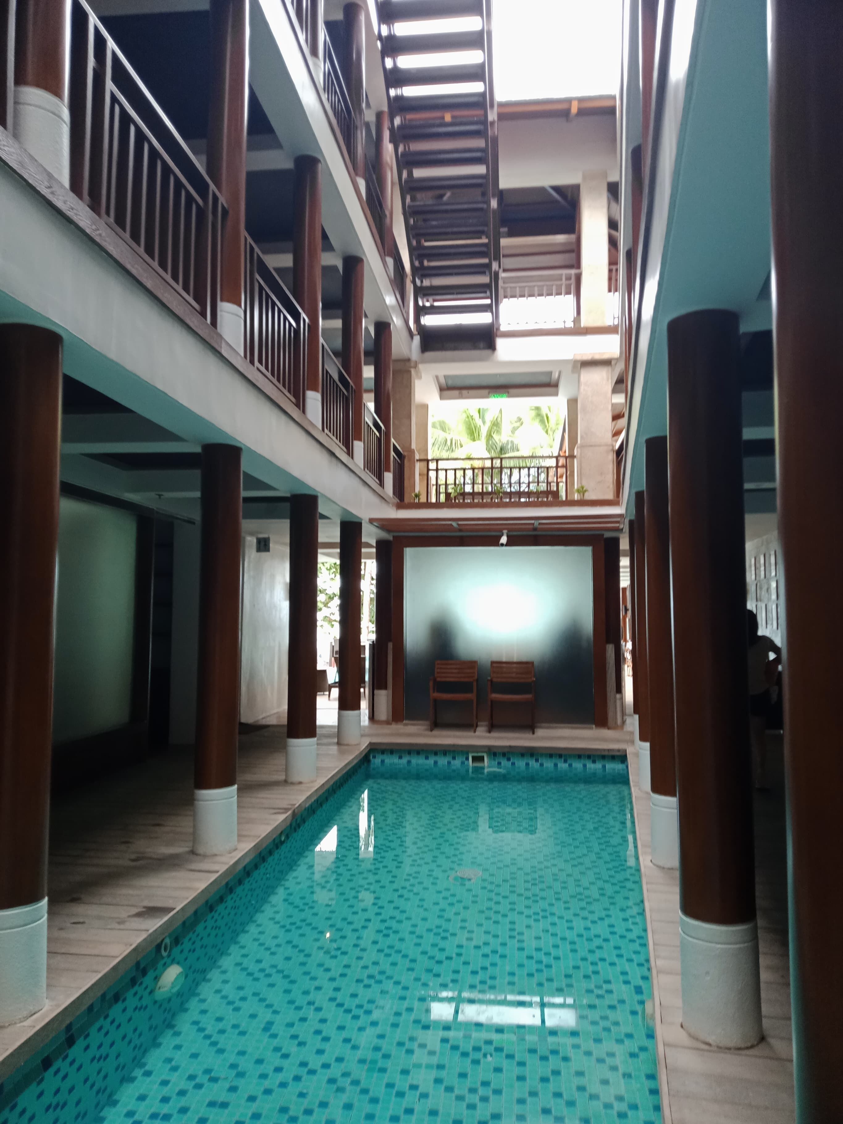 An indoor swimming pool with tiled flooring, wooden pillars, and a balcony with railings.
