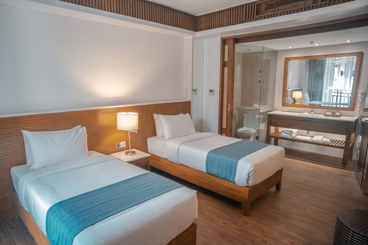 Habitación de hotel con dos camas, un cabecero de madera, una mesita de noche con lámpara, un baño con espejo y suelo de madera.