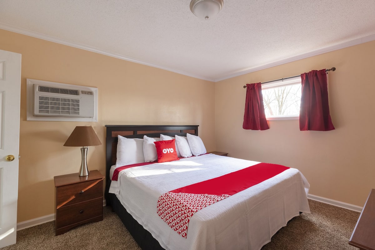 A hotel room with a bed, a nightstand, a lamp, an air conditioning unit, a window with curtains, and a door.