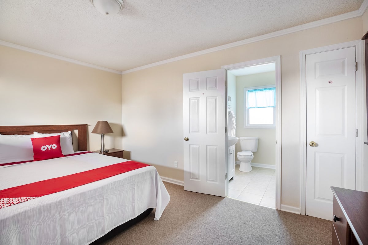 A hotel room with a bed, a nightstand, a lamp, a bathroom visible through an open door, and a wardrobe.