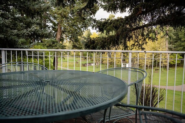 Une table et des chaises en métal sur un balcon doté d’une balustrade blanche, avec vue sur une pelouse bien entretenue et des arbres.