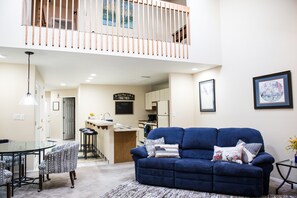 A spacious living area with a blue sofa, a glass dining table, and a kitchen in the background.