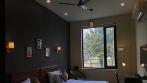 A hotel room with two beds, a desk, a chair, a ceiling fan, and a window with a view of trees. - Ashira Inn (Greater Noida)