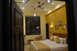 A hotel room with a bed, a desk, a chair, a window, and a ceiling fan. - Ashira Inn (Greater Noida)