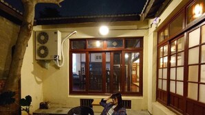 Una persona sentada en una silla en una habitación con piso de baldosas, una ventana con marcos de madera y un aire acondicionado instalado en la pared.