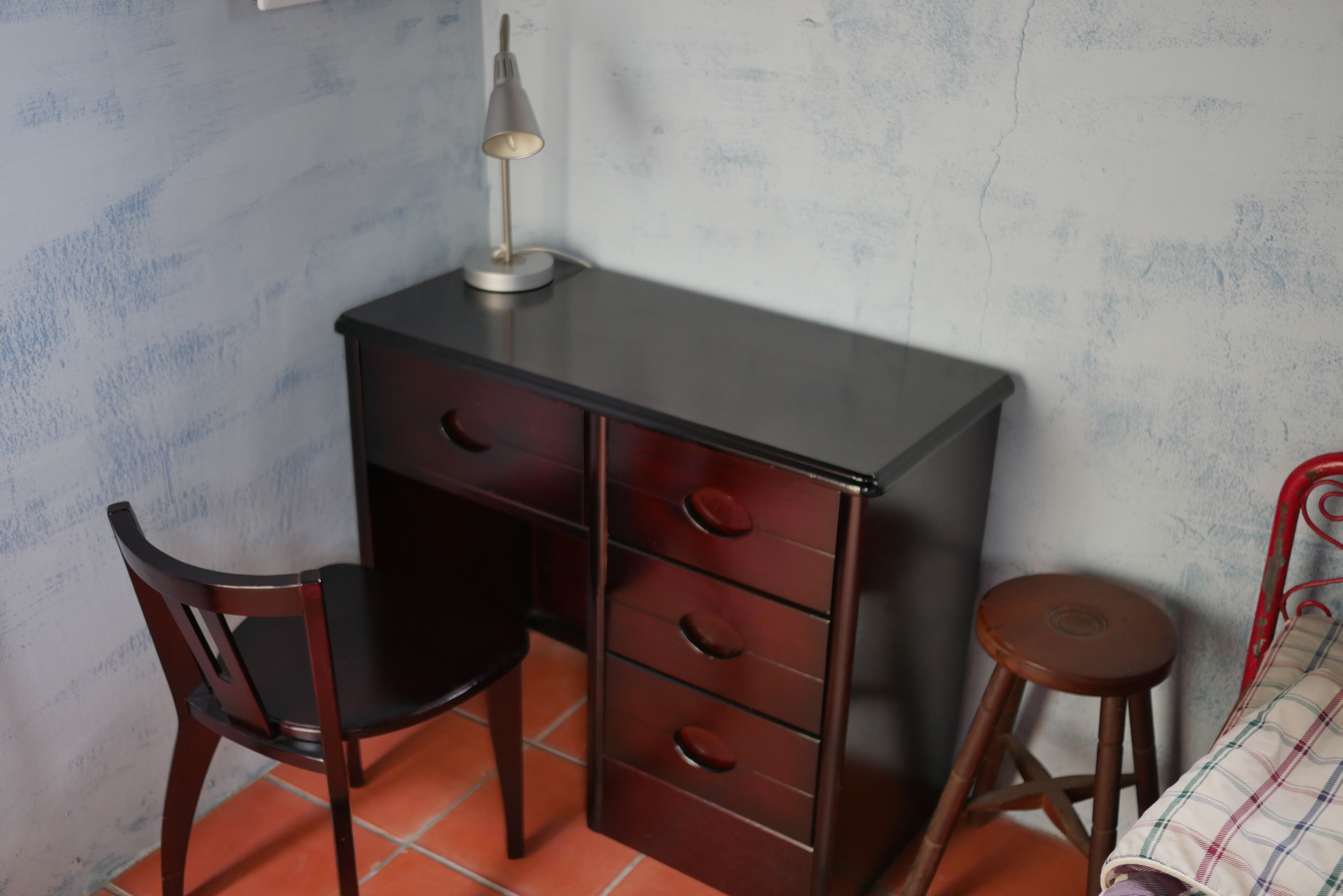 A small room with a desk, chair, and stool. There is a lamp on the desk.