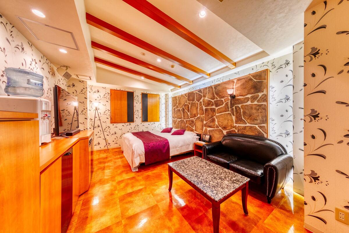 A room with a bed, a sofa, a coffee table, a desk with a water dispenser, and a stone wall.