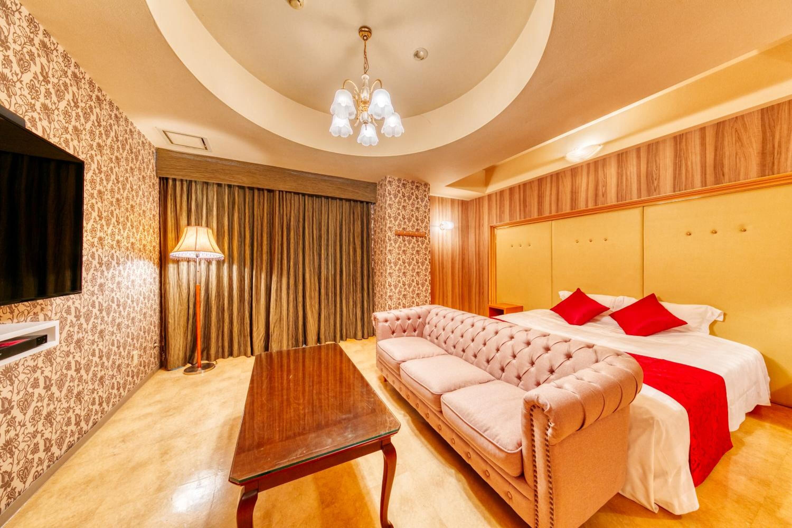 A hotel room with a tufted sofa, a bed with red pillows, a wooden headboard, a flat-screen TV, and a chandelier.