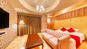 A hotel room with a tufted sofa, a bed with red pillows, a wooden headboard, a flat-screen TV, and a chandelier.