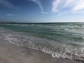A beach with waves gently lapping onto the shore, a clear sky, and a distant shoreline with buildings.