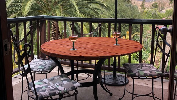 Et træbord med to stole, en orange parasol og et glas med drik på en balkon med udsigt til palmer og bakker.
