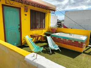 A small outdoor area with a yellow wall, a turquoise door, a window with bars, a small table, two chairs, and a bathtub with tiled walls.