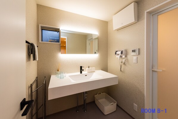 Une salle de bain moderne avec un lavabo blanc, un miroir et un porte-serviettes. Il y a une petite fenêtre au-dessus du miroir, un panier sous le lavabo et une porte sur la droite.