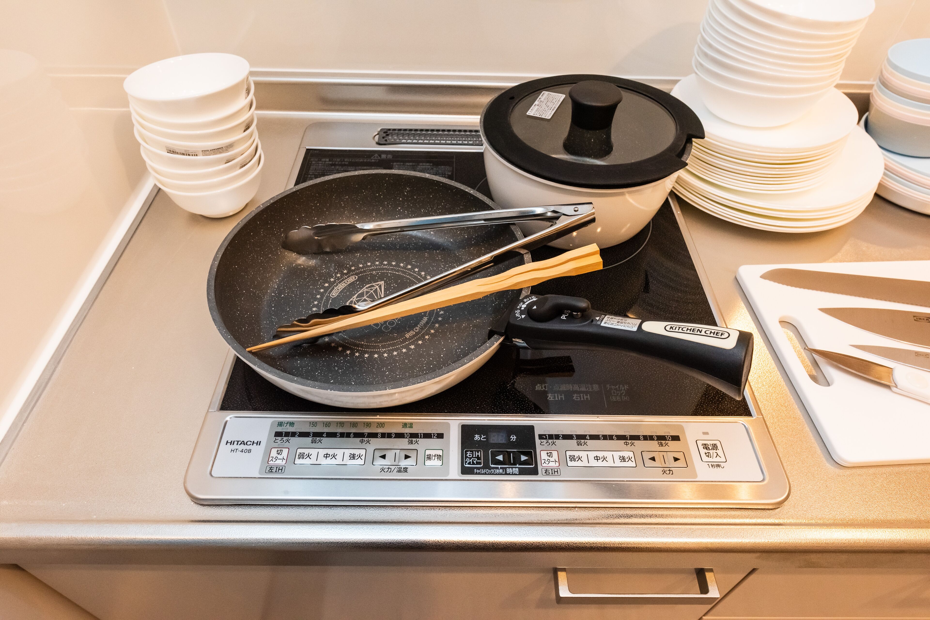 A kitchen with a stove, a frying pan, tongs, a pot, and stacked plates.