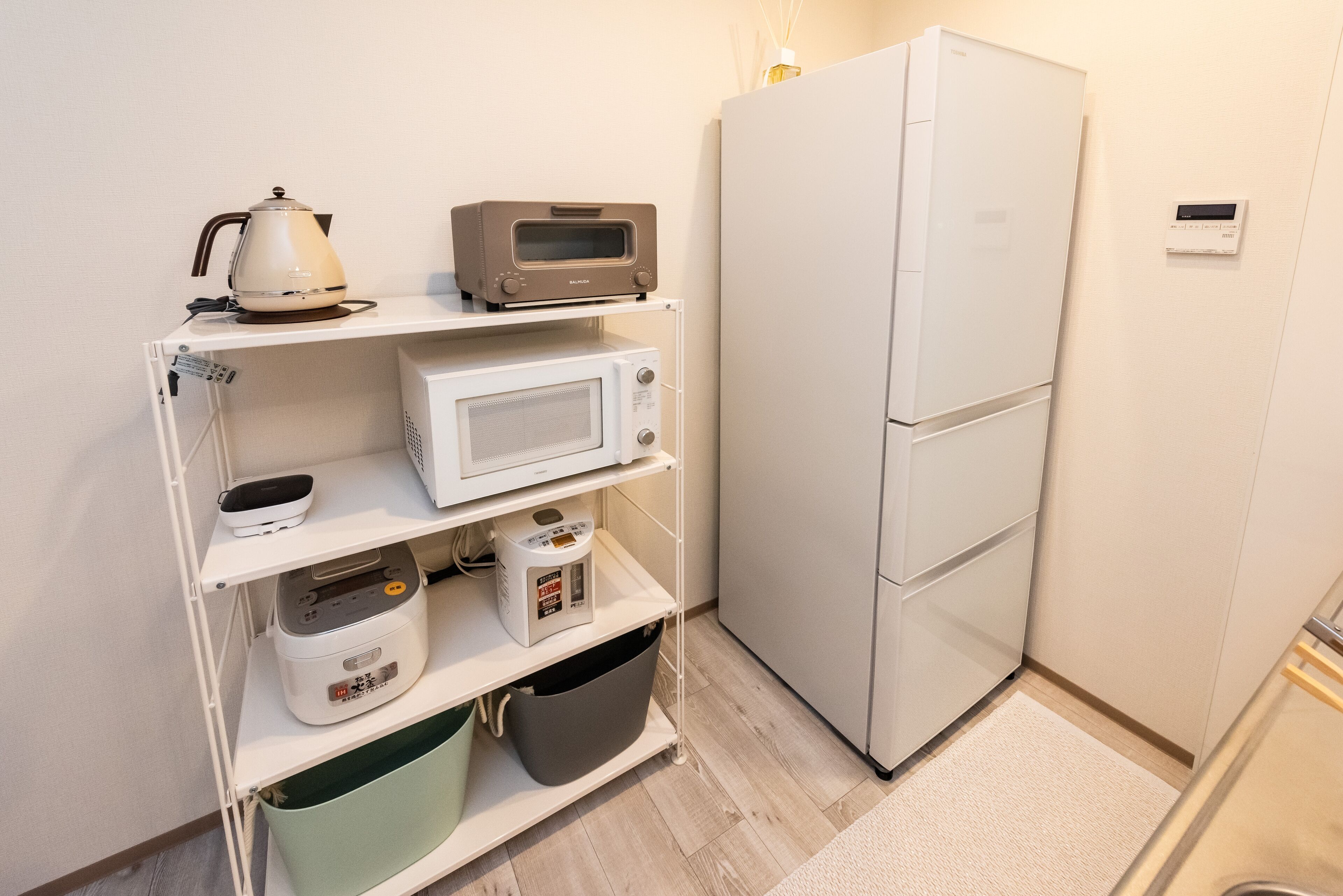 A compact kitchenette with a microwave, toaster, and kettle on a shelf, a refrigerator, and a trash bin.