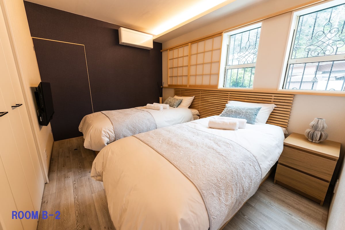 Un dormitorio con dos camas, una mesita de noche, un aire acondicionado de pared y una ventana con herrajes decorativos.