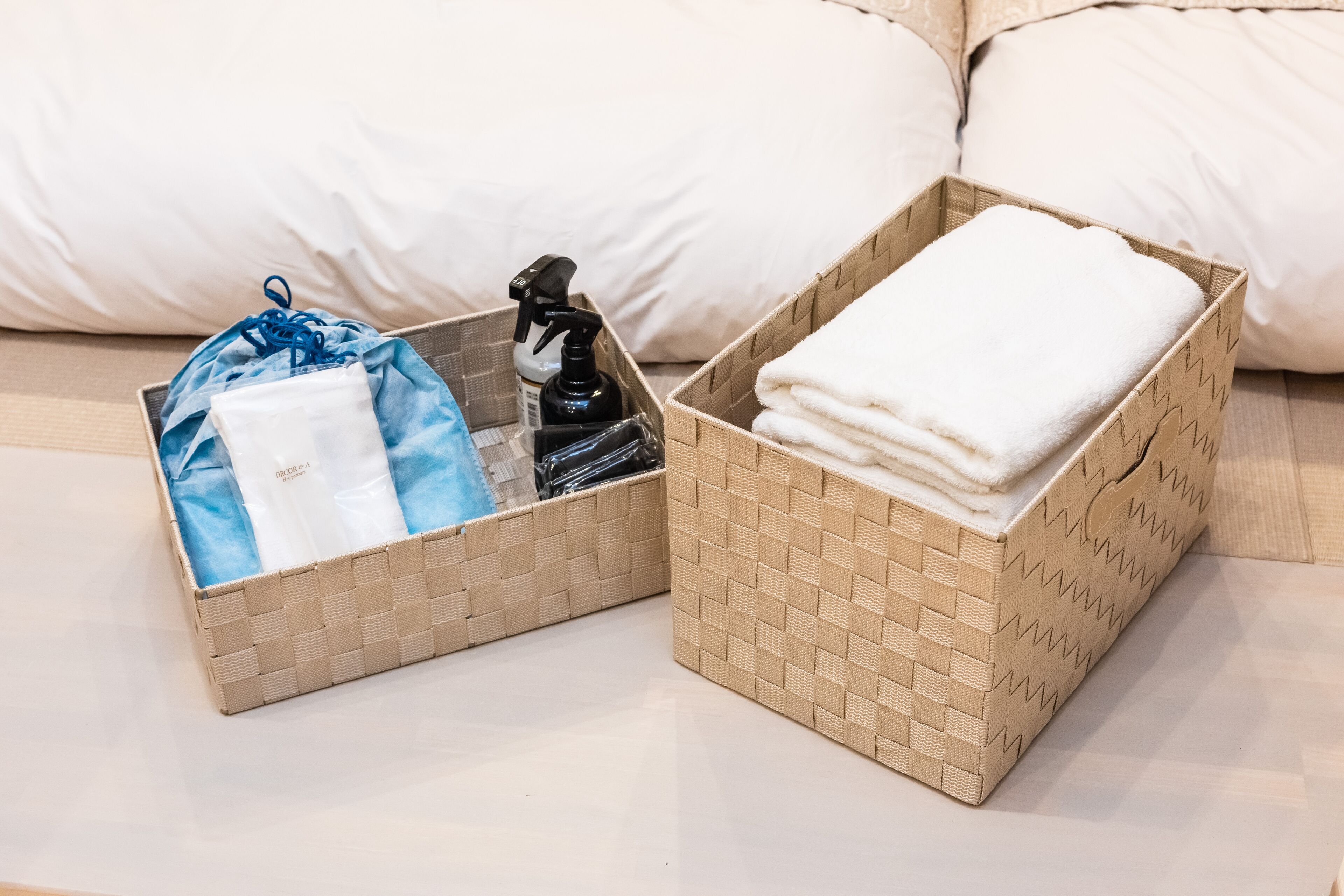 Two wicker baskets on a bed, one with a blue bag and a spray bottle, the other with folded white towels.