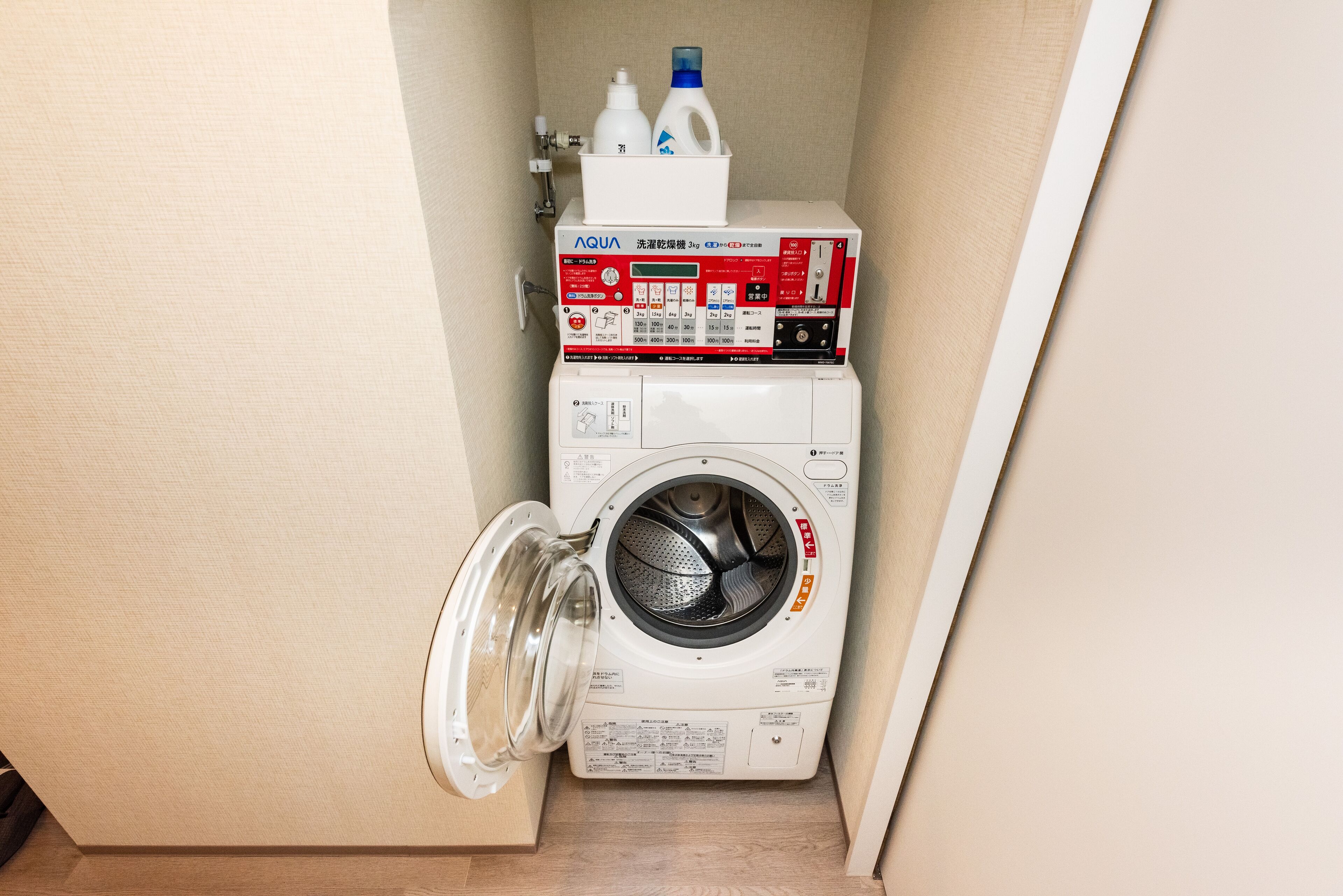A compact washing machine in a small laundry room with a control panel and detergent bottles on top.