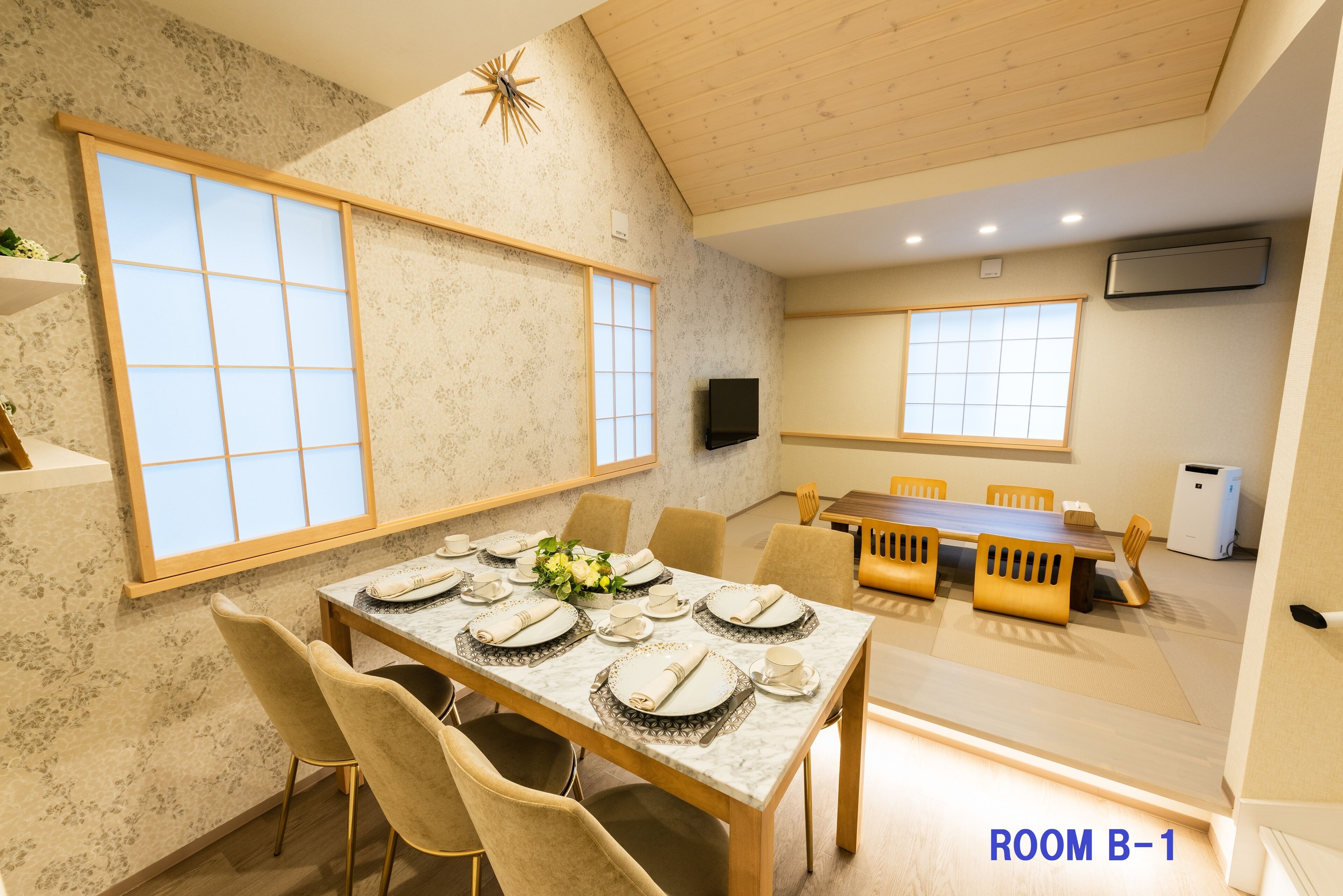 A dining area with a wooden table set for six, beige chairs, and large windows with wooden frames.