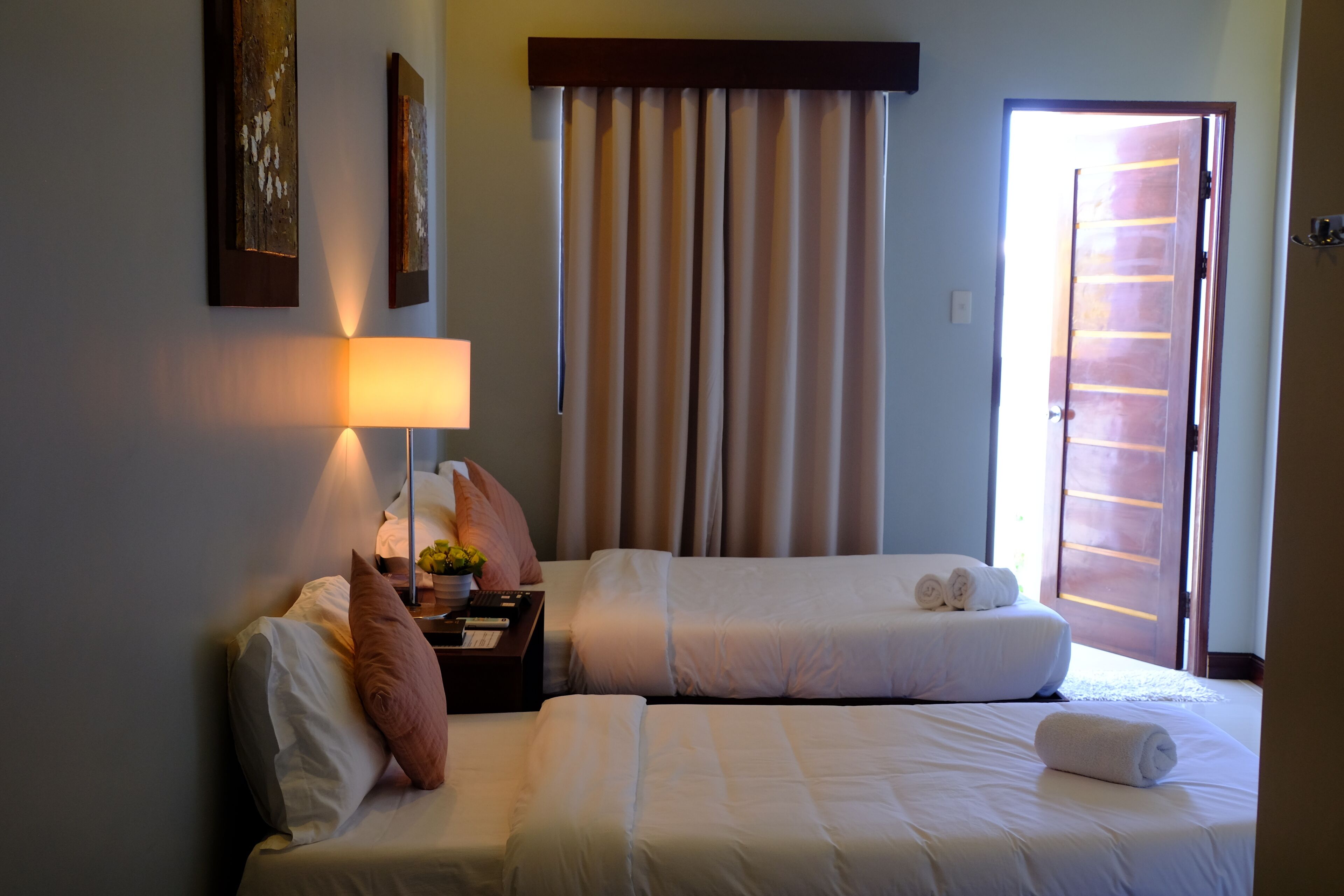 Habitación de hotel con dos camas, una mesita de noche con lámpara, una ventana con cortinas y una obra de arte en la pared.