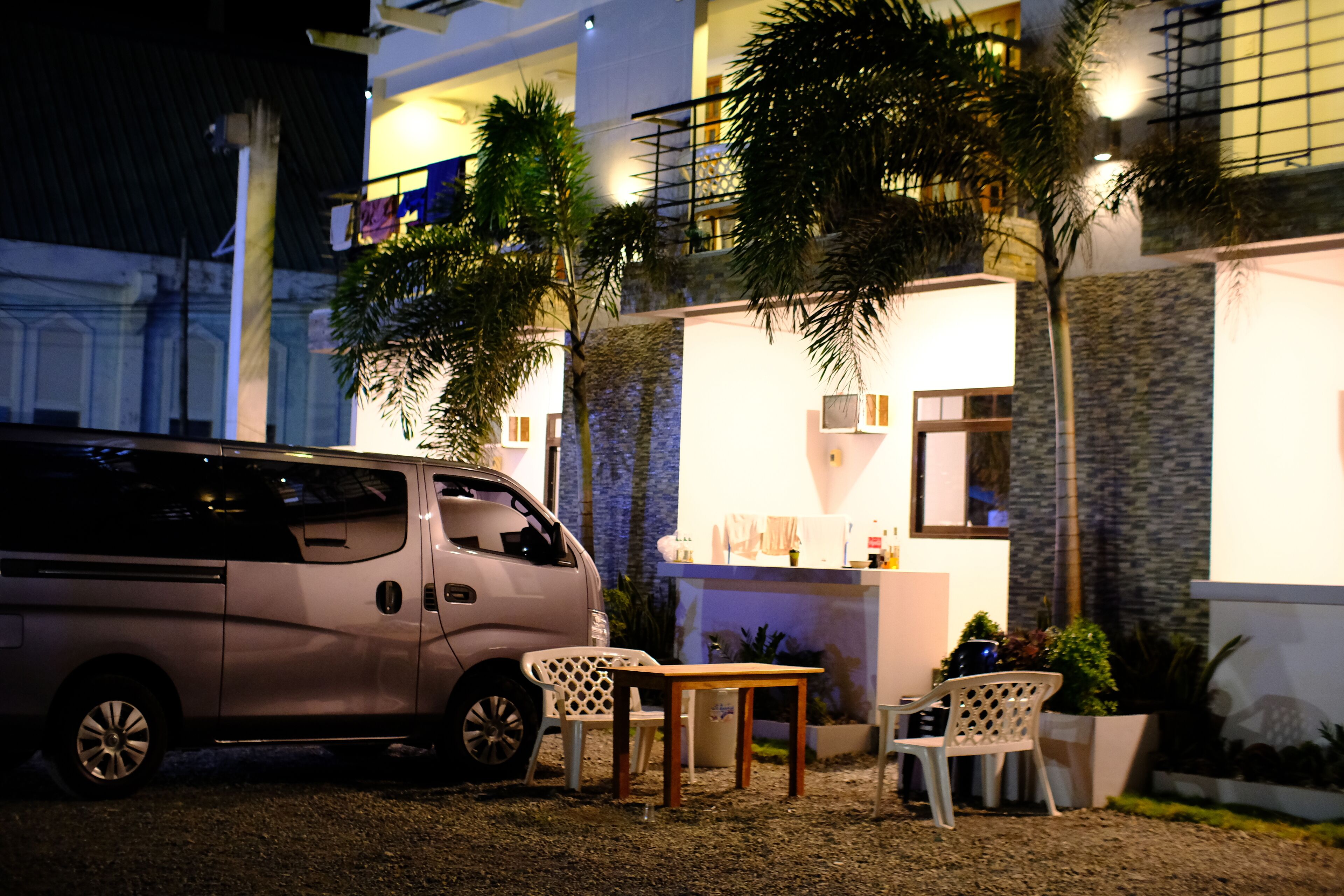 Una camioneta estacionada frente a un edificio con un patio, que incluye una mesa y sillas, y un pequeño jardín con plantas.