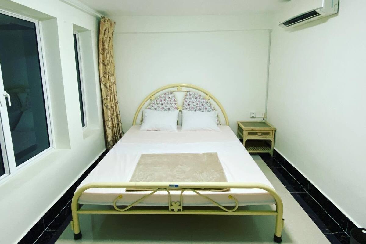 A single-bed room with a headboard, a bedside table, a window with curtains, and an air conditioning unit on the wall.