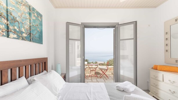 A bedroom with a bed, a dresser, a painting, and a view of the sea through an open door. - Villa Bianca Tzia (Kea)