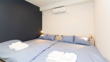 A hotel room with two beds, each with blue bedding and white towels. There is a wall-mounted air conditioner above the beds.