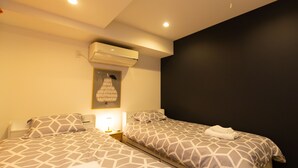 A hotel room with two beds, each with a geometric patterned bedspread, a wall-mounted air conditioner, a bedside lamp, and a framed artwork above the left bed. - Cross Bridge Ikebukuro (Tokyo)