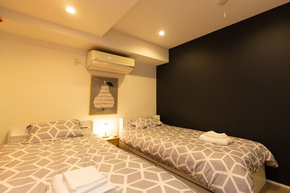 Una habitación de hotel con dos camas, cada una con una colcha de estampado geométrico, aire acondicionado montado en la pared, una lámpara de noche y una obra de arte enmarcada sobre la cama izquierda.