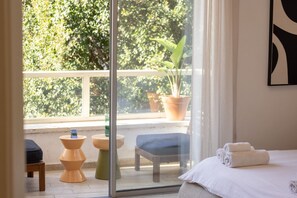 A hotel room with a balcony, a bed with white linens, a small table with a blue chair, and potted plants.