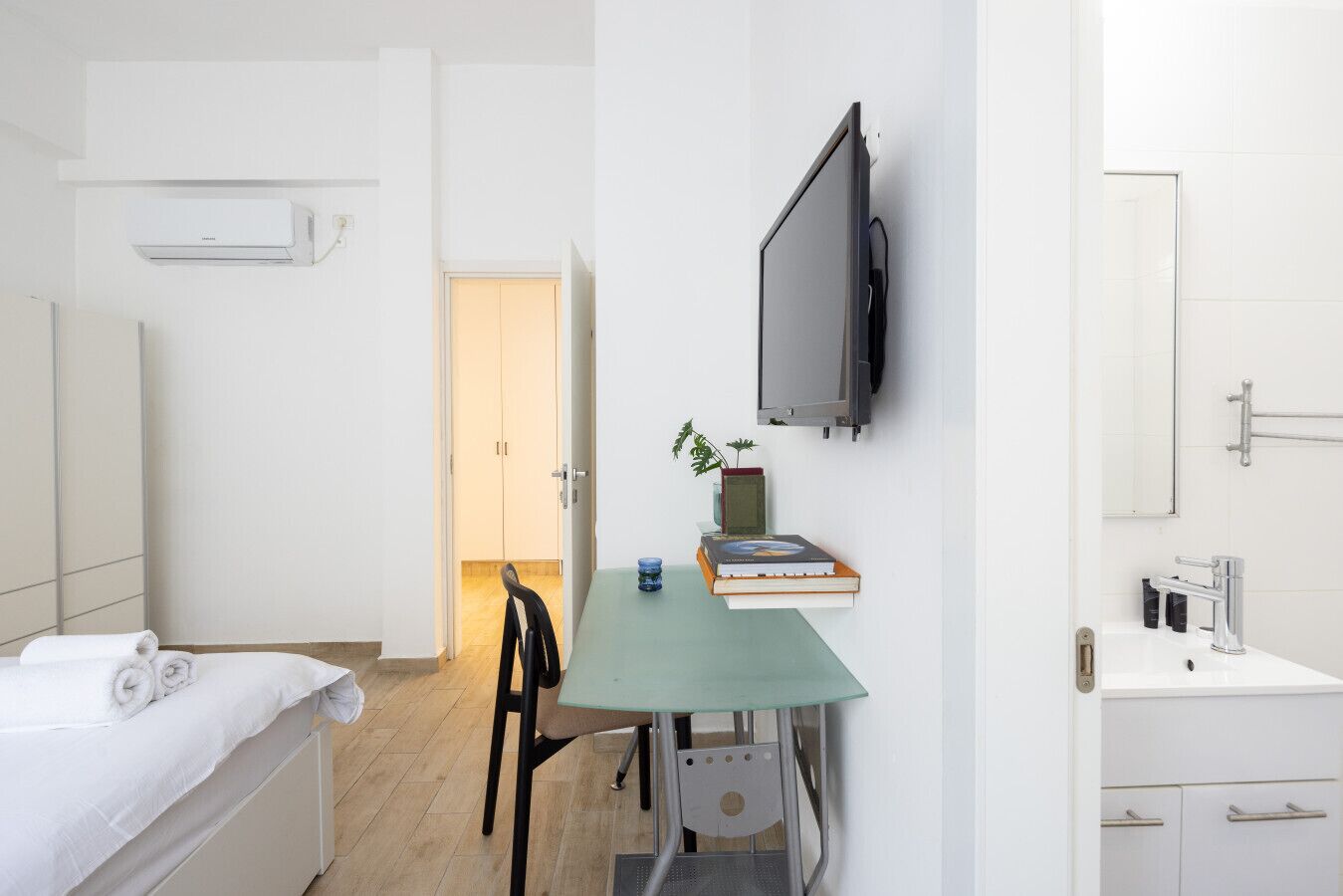 Habitación de hotel con cama, escritorio con silla, televisor montado en la pared y baño con lavamanos y espejo.