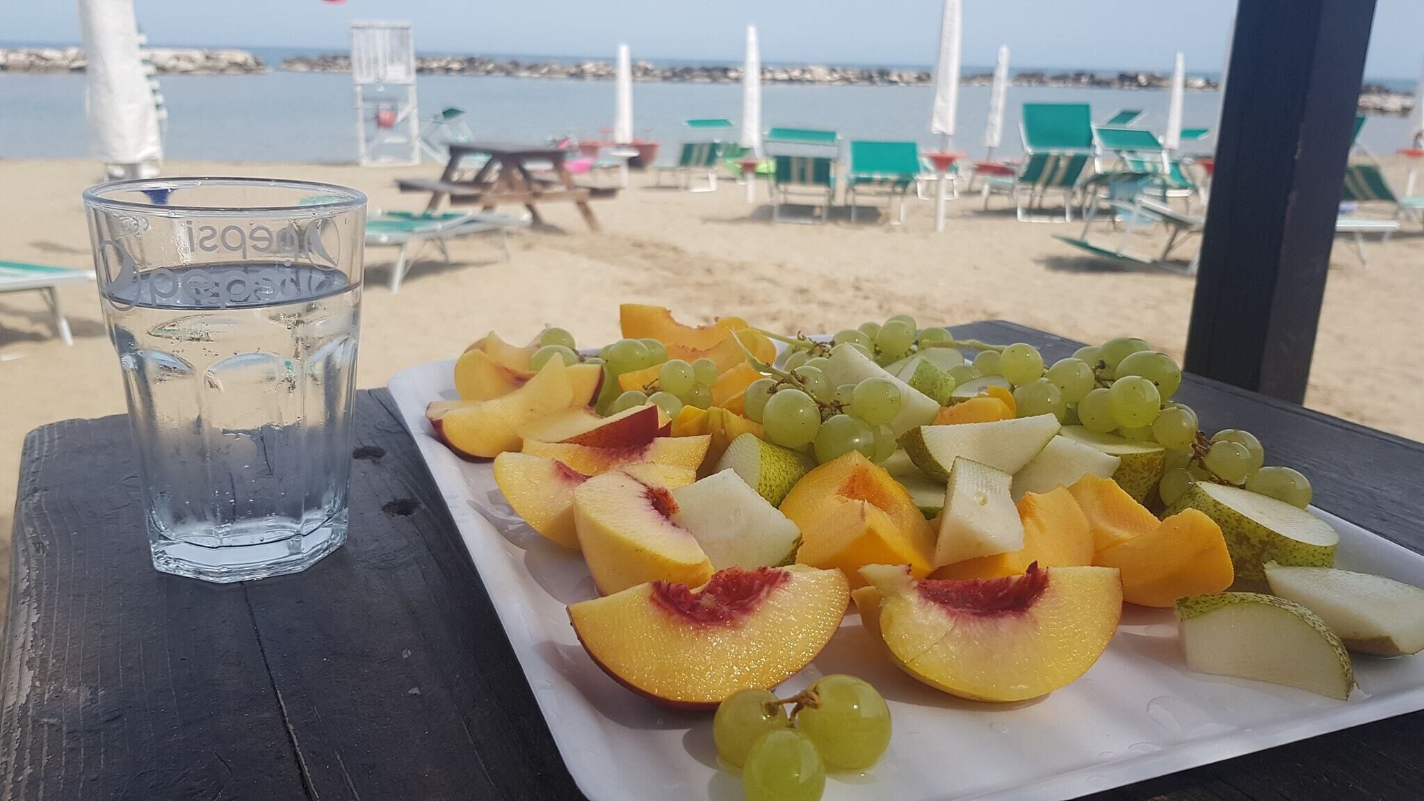 A table with a plate of fruit and a glass of water on a beach.
