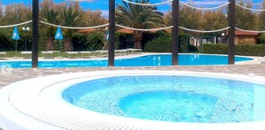 En rund pool med klart, blåt vand, omgivet af liggestole og palmer.